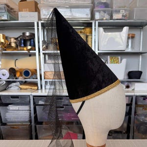 A black velvet hat with a gold trim, placed on a mannequin head in a workshop filled with various crafting supplies and materials.