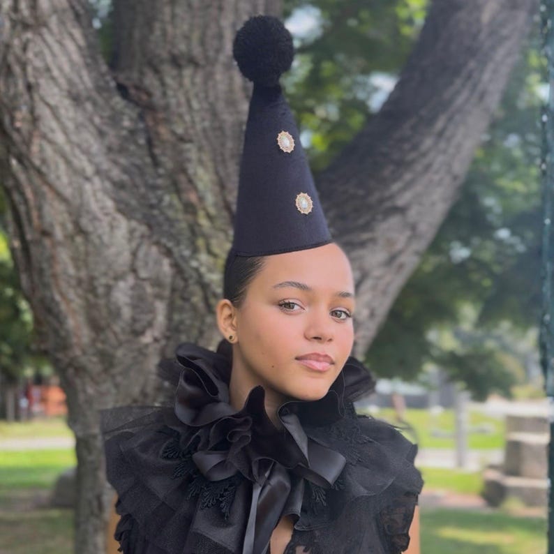 A woman wearing a black party hat with a pom pom and a black ruffled collar stands in front of a tree.