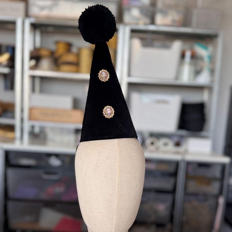A black pom pom hat with two gold embellishments is displayed on a mannequin head in a room with shelves and other items in the background.
