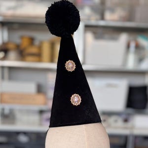 A black pom pom hat with two gold-colored embellishments on the front, displayed on a mannequin head.