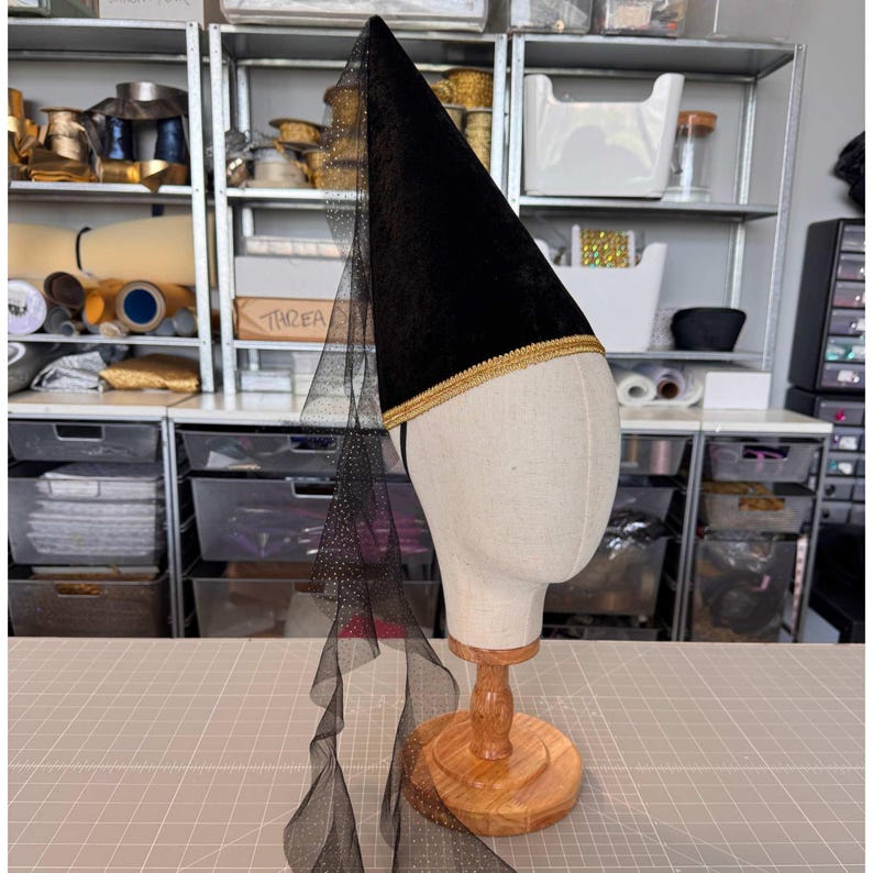 a mannequin head wearing a black velvet hat with a gold trim, with a long, flowing veil draped over it, placed on a wooden stand.