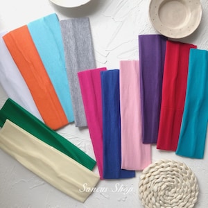 May include: Assortment of 12 colourful fabric headbands in various colours, including white, orange, light blue, grey, pink, blue, purple, red, and turquoise. The headbands are arranged in a fan shape on a white surface. Sancus Shop is written in black text on the bottom of the image.