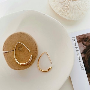 May include: A pair of gold-toned hoop earrings with a unique teardrop shape. The earrings are displayed on a wooden surface and a white plate.