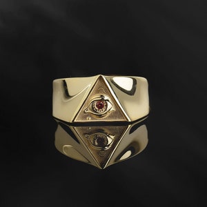 Puede incluir: Un anillo de oro con un dise&ntilde;o triangular que presenta un ojo con una piedra preciosa roja en el centro.