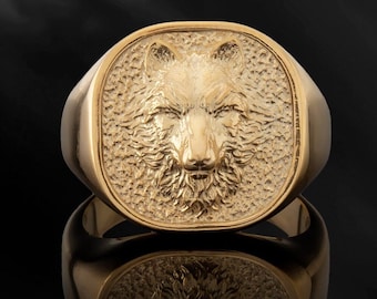 May include: A gold signet ring with a detailed wolf head design. The ring is square shaped and has a textured surface.