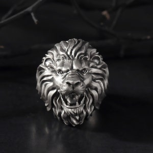 Puede incluir: Un anillo de cabeza de le&oacute;n de plata con un dise&ntilde;o detallado. La boca del le&oacute;n est&aacute; abierta y sus dientes son visibles. El anillo est&aacute; hecho de metal y tiene un acabado brillante.