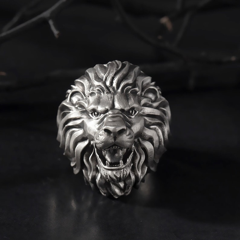 Puede incluir: Un anillo de cabeza de le&oacute;n de plata con un dise&ntilde;o detallado. La boca del le&oacute;n est&aacute; abierta y sus dientes son visibles. El anillo est&aacute; hecho de metal y tiene un acabado brillante.