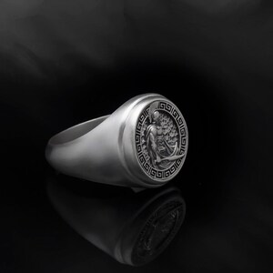 May include: A silver ring with a raised circular design featuring a Greek warrior holding a sword and shield. The design is surrounded by a Greek key border.