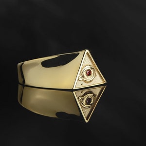 Puede incluir: Un anillo de oro con un dise&ntilde;o de pir&aacute;mide que presenta un ojo con una piedra preciosa roja. El anillo se refleja en una superficie negra.