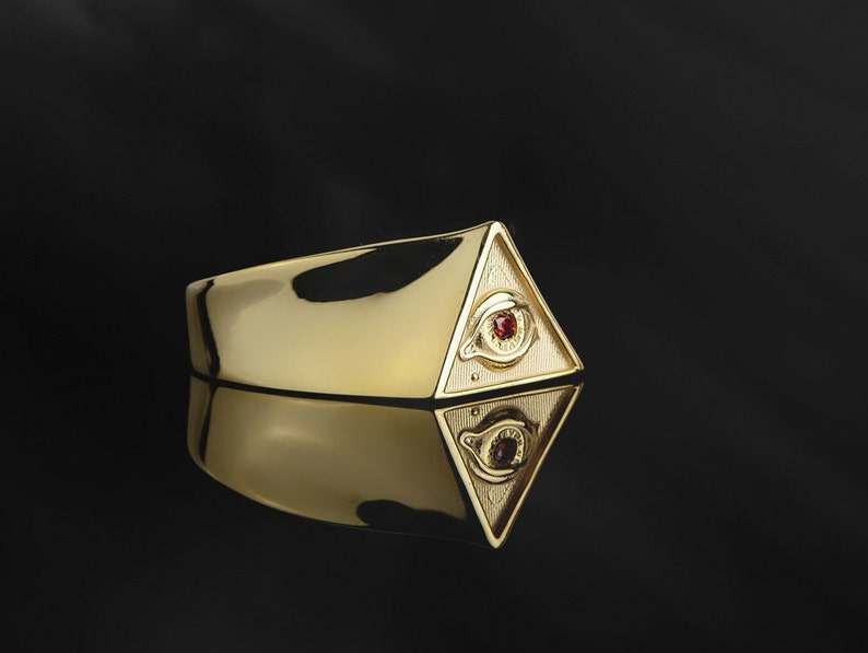 Puede incluir: Un anillo de oro con un dise&ntilde;o de pir&aacute;mide que presenta un ojo con una piedra preciosa roja. El anillo se refleja en una superficie negra.