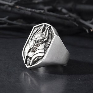 May include: A silver ring with a detailed design of a creature's head with a skull underneath. The ring is set in a shield-shaped setting.