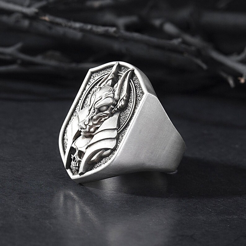 May include: A silver ring with a detailed design of a creature's head with a skull underneath. The ring is set in a shield-shaped setting.