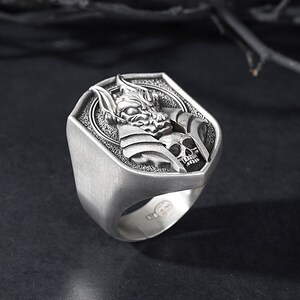 May include: A silver ring with a detailed design of a creature with a skull. The ring is shaped like a shield.