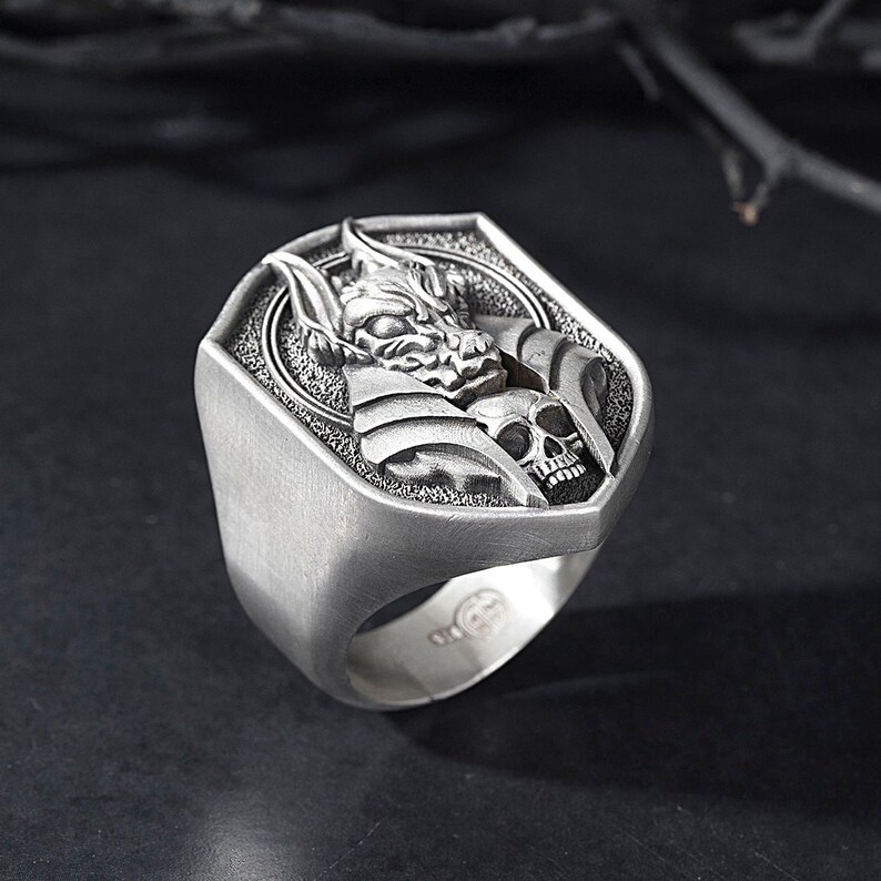 May include: A silver ring with a detailed design of a creature with a skull. The ring is shaped like a shield.