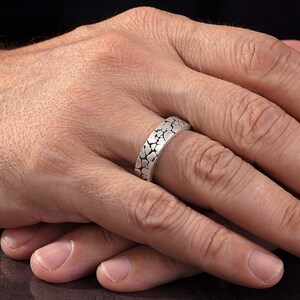 May include: A silver ring with a textured design. The ring is worn on a person's finger.