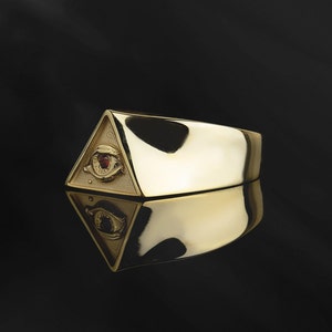 Puede incluir: Un anillo de oro con un dise&ntilde;o triangular que presenta un ojo que todo lo ve con una piedra preciosa roja en el centro.
