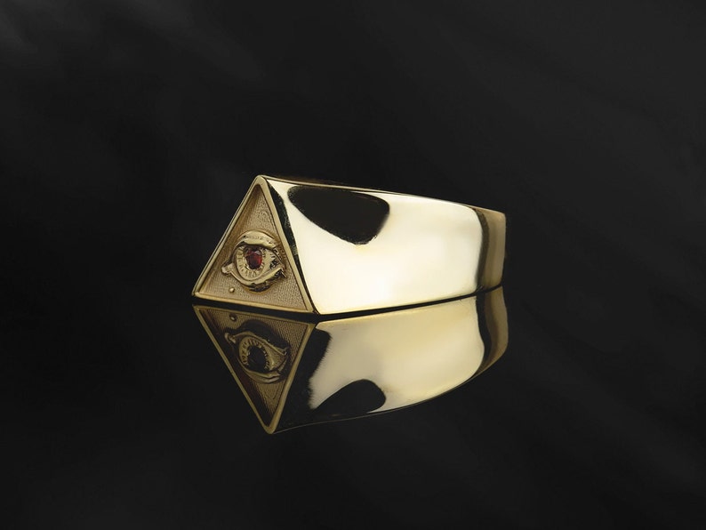 Puede incluir: Un anillo de oro con un dise&ntilde;o triangular que presenta un ojo que todo lo ve con una piedra preciosa roja en el centro.