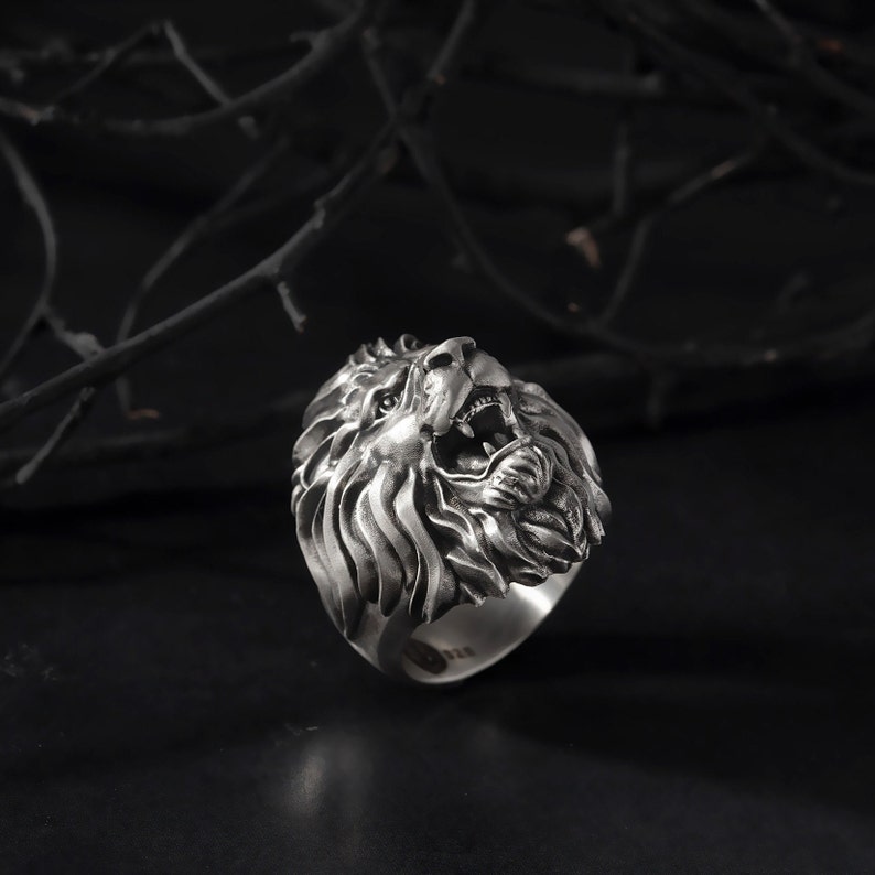 Puede incluir: Un anillo de plata con un dise&ntilde;o detallado de cabeza de le&oacute;n. La boca del le&oacute;n est&aacute; abierta y su melena est&aacute; esculpida de forma intrincada.