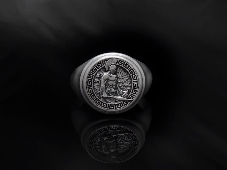 May include: A silver ring featuring a detailed design of a Greek warrior in a helmet and armour, surrounded by a Greek key pattern.