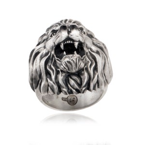 Puede incluir: Un anillo de plata con un dise&ntilde;o detallado de cabeza de le&oacute;n. La boca del le&oacute;n est&aacute; abierta y sus dientes son visibles. El anillo est&aacute; hecho de plata de ley y tiene un sello en la banda.
