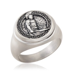 May include: A silver ring with a round, raised design featuring a Greek warrior holding a sword. The design is surrounded by a black Greek key border.
