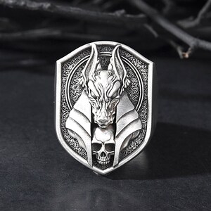 May include: A silver ring with a detailed design of an Anubis head with a skull underneath. The ring is in the shape of a shield.