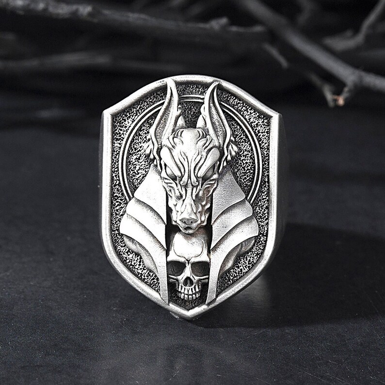 May include: A silver ring with a detailed design of an Anubis head with a skull underneath. The ring is in the shape of a shield.