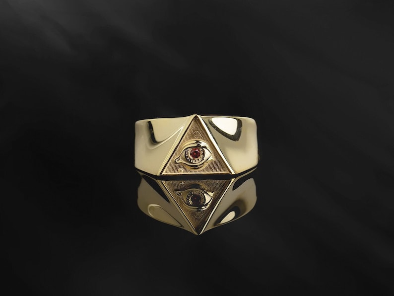 Puede incluir: Un anillo de oro con un dise&ntilde;o triangular que presenta un ojo con una piedra preciosa roja en el centro.