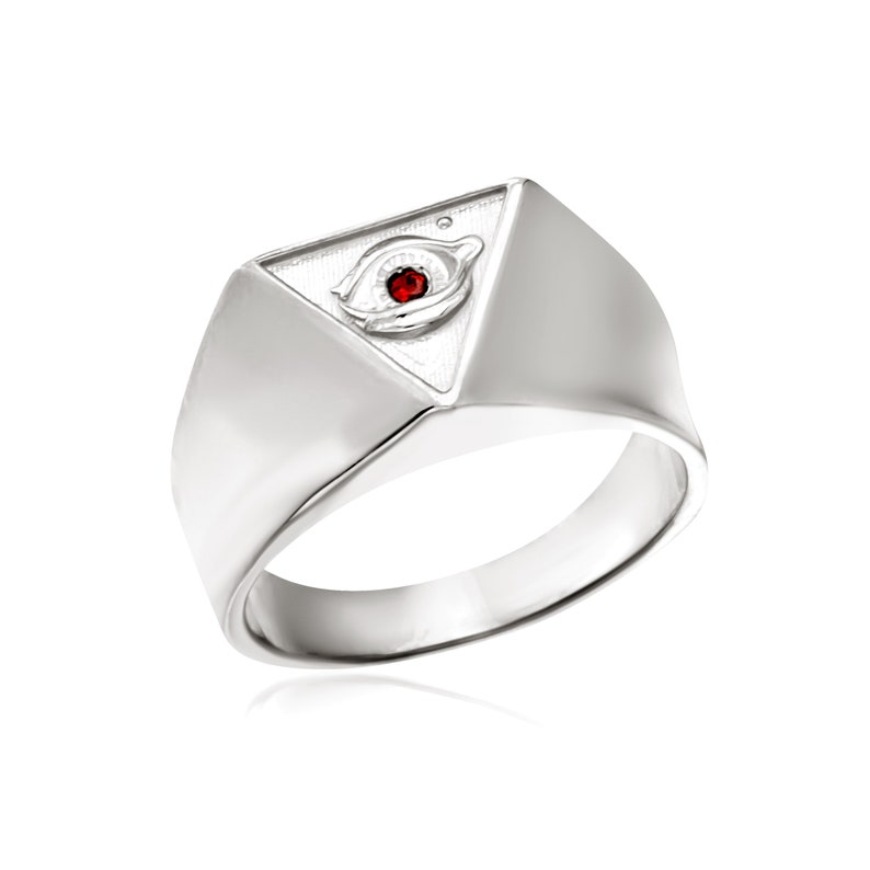 Puede incluir: Un anillo de plata con un dise&ntilde;o triangular que presenta un ojo con una piedra preciosa roja en el centro.