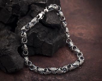 May include: A silver chain bracelet with a detailed, intricate design. The chain is made of small, round links with a decorative pattern. The bracelet is lying on a dark, textured surface.