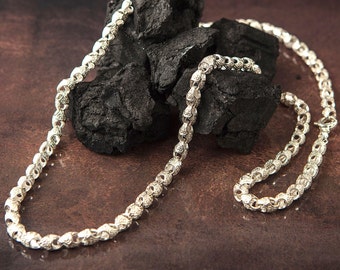 May include: A silver chain necklace with a detailed, ornate design. The necklace is displayed on a bed of black charcoal.