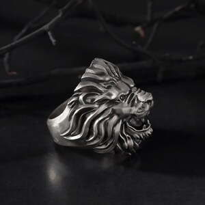 Puede incluir: Un anillo de cabeza de le&oacute;n de plata con un dise&ntilde;o detallado. El anillo est&aacute; hecho de metal y tiene una superficie texturizada.