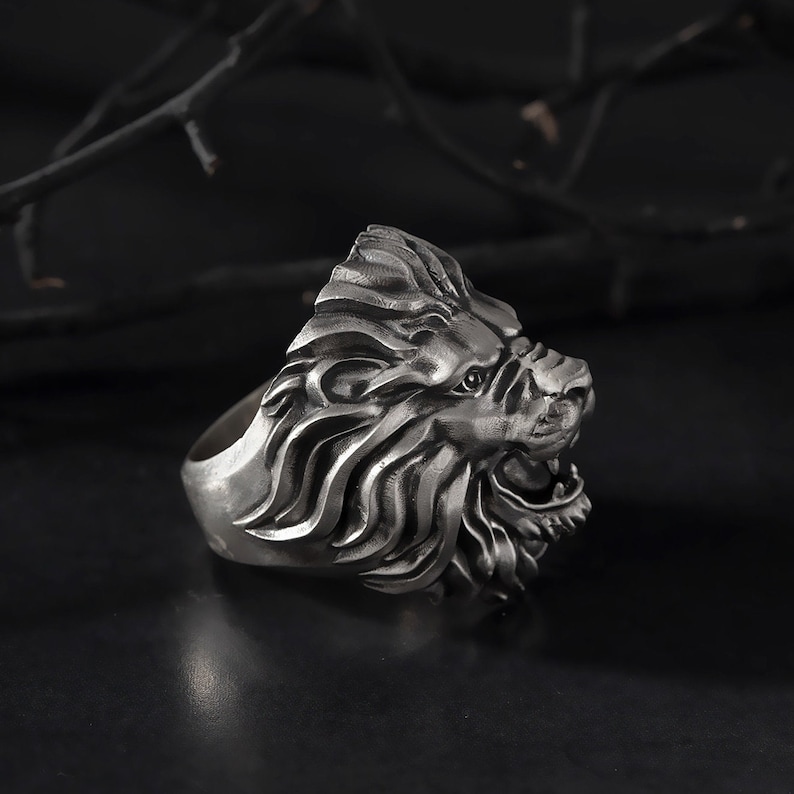 Puede incluir: Un anillo de cabeza de le&oacute;n de plata con un dise&ntilde;o detallado. El anillo est&aacute; hecho de metal y tiene una superficie texturizada.