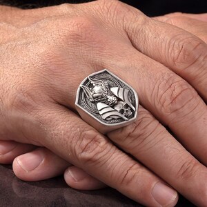 May include: A silver ring with a detailed design of a wolf's head and a skull. The ring is set in a shield-shaped setting.
