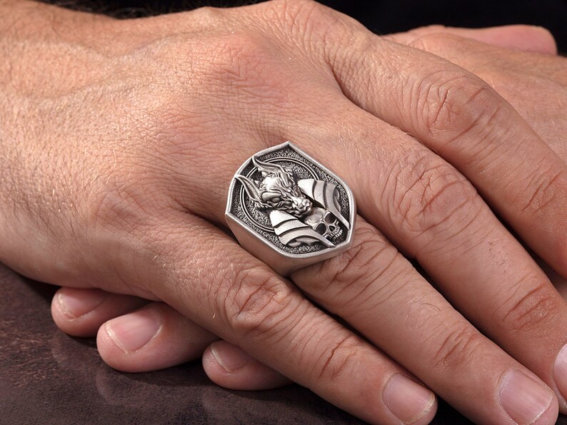 May include: A silver ring with a detailed design of a wolf's head and a skull. The ring is set in a shield-shaped setting.