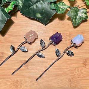 May include: Three gemstone rose hair sticks. Each rose is a different color: pink, purple, and white. The roses are mounted on silver-colored stems with leaves. The background is a light brown wooden surface.