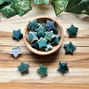 May include: A bowl filled with green and white agate star-shaped stones. The bowl is made of wood and has a natural finish. The stones are arranged in a random pattern. There are also several star-shaped stones scattered around the bowl on a wooden surface.