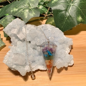 May include: A clear crystal pendulum with a silver chain and a brown bead at the end. The pendulum is filled with colorful crystals and stones, including amethyst, turquoise, and carnelian.