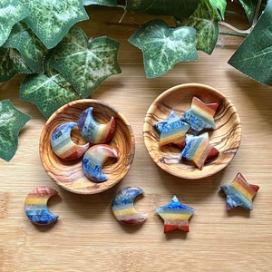May include: Seven rainbow chakra stones, four shaped as crescent moons and three shaped as stars, are displayed in two small wooden bowls. The stones are made of layered semi-precious stones in shades of red, orange, yellow, green, blue, indigo, and violet.