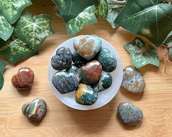 May include: A collection of heart-shaped, polished, green, brown, and red jasper stones in a white bowl. The stones are arranged in a circle, with the points of the hearts facing outward.