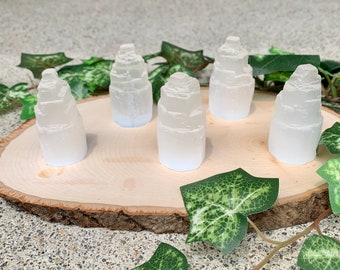 Peut inclure: Cinq tours de cristal de sélénite blanche sur une tranche de bois avec des feuilles vertes. Les cristaux sont empilés en forme de pyramide et ont une surface lisse et polie.