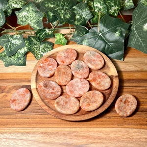 May include: A collection of 12 sunstone palm stones in various shades of orange and white. The stones are arranged on a wooden plate.