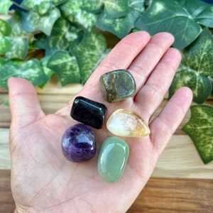 May include: Five tumbled stones in various colors, including green, black, purple, yellow, and brown. The stones are arranged in a hand, with the green stone in the foreground.