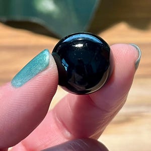 May include: A polished, spherical black gemstone, reflecting light. The smooth, glossy surface of the sphere is held between fingers with teal nail polish. The background is a blurred view of wood and green foliage.