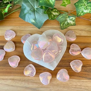 May include: A collection of pink heart-shaped crystals with iridescent shimmer. The crystals are arranged in a white heart-shaped dish and on a wooden surface.