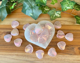 May include: A collection of pink heart-shaped crystals with iridescent shimmer. The crystals are arranged in a white heart-shaped dish and on a wooden surface.