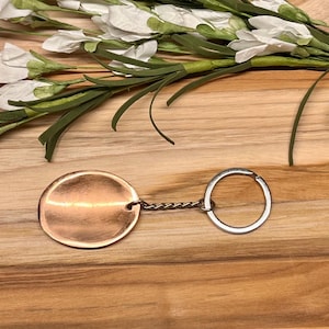 May include: A circular copper-toned keychain featuring a smooth, reflective surface. The metal disc is attached to a standard silver-colored split ring by a short, twisted metal chain, creating a simple and functional accessory for keys.