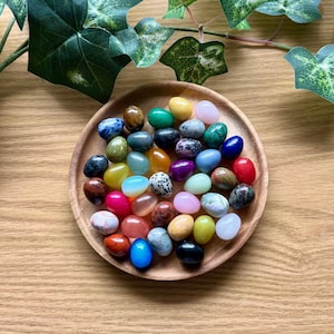 May include: A wooden bowl filled with a variety of colorful, smooth, polished stones in the shape of eggs. The stones are arranged in a random pattern and are all different sizes and shapes.