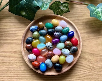 May include: A wooden bowl filled with a variety of colorful, smooth, polished stones in the shape of eggs. The stones are arranged in a random pattern and are all different sizes and shapes.