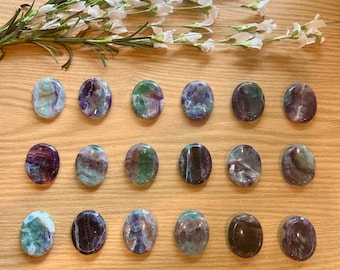 May include: A collection of oval-shaped fluorite stones in shades of purple, green, and blue, arranged on a light wood surface. The stones have a polished, smooth texture. White flowers and green stems are in the background.